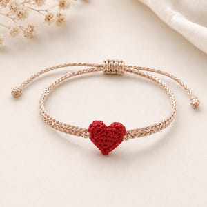Puede incluir: Una pulsera de macramé beige con un dije en forma de corazón rojo. La pulsera ajustable presenta un diseño tejido y un pequeño corazón de ganchillo en el centro. La pulsera se muestra sobre una superficie blanca.