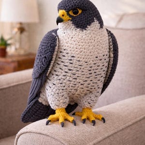 Pode incluir: Escultura de falcão de crochê com cabeça e asas cinza escuro, corpo creme e pés amarelos. O pássaro tem um bico amarelo e garras pretas. Está pousado em uma superfície estofada bege.