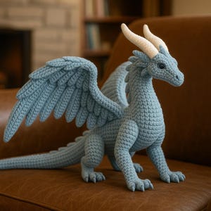 Op de afbeelding: Een lichtblauwe gehaakte draak met grote vleugels en crèmekleurige hoorns. De draak staat op een bruin leren oppervlak. De vleugels van de draak zijn gespreid en zijn staart is uitgestrekt.