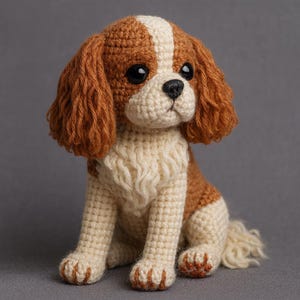 May include: A crocheted Cavalier King Charles Spaniel dog toy. The handmade plush dog is in a sitting pose, with a white and brown colour scheme. It has large black eyes, a black nose, and brown ears and markings. The paws are detailed.