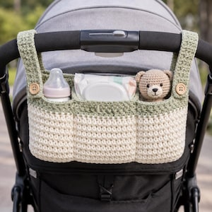 Könnte beinhalten: Ein gehäkelter Kinderwagen-Organizer in Creme und Salbeigrün, befestigt an einem Kinderwagen-Griff. Der Organizer hat drei Fächer mit einer Babyflasche, Feuchttüchern und einem kleinen Teddybären. Holzknöpfe befestigen den Organizer am Kinderwagen.