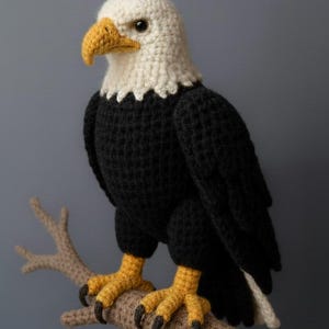 Puede incluir: Un águila calva de ganchillo, con cabeza blanca y pico amarillo, posada en una rama. El cuerpo del águila es negro, con patas y garras amarillas. La rama es de color marrón claro, sobre un fondo gris. Un artículo decorativo hecho a mano.