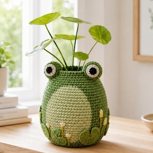 May include: A handcrafted, green crochet frog planter holding a leafy green plant. The frog planter has large, round eyes and a light green belly. Lily pads and small white flowers decorate the base of the planter. The planter sits on a wooden surface.