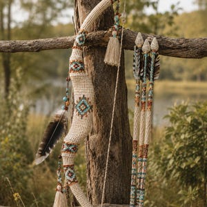 May include: A handcrafted, off-white crochet bow and three arrows with decorative patterns in brown, turquoise, and beige. The bow features a feather and tassels, while the arrows have feathered fletching. The set is displayed outdoors.