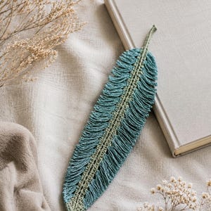May include: A handmade, teal and green crochet feather bookmark. The bookmark is lying on a neutral-colored surface next to a closed book. The feather design has a detailed fringe and a central, lighter green spine.