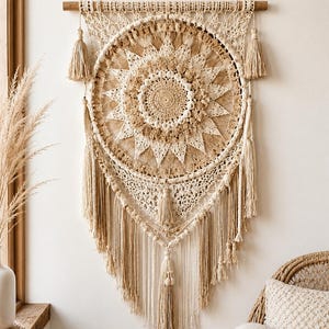 May include: A large, beige macrame wall hanging with a circular design. The central circle features intricate crochet work in varying shades of beige and tan. Long tassels hang from the bottom, and smaller tassels are on the sides. The wall hanging is suspended from a wooden rod.