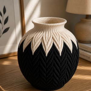 May include: A handcrafted, decorative vase with a unique design. The vase features a cream-colored top and leaf-like accents, transitioning to a black, chevron-patterned base. The vase is made of a soft, textured material and is suitable for home decor.