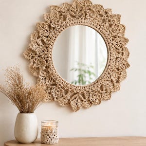 May include: A circular mirror with a crocheted border in a beige hue, adorned with floral and leaf motifs. The mirror hangs on a light wall. A vase of dried flowers and a candle in a crocheted sleeve sit on a wooden table.