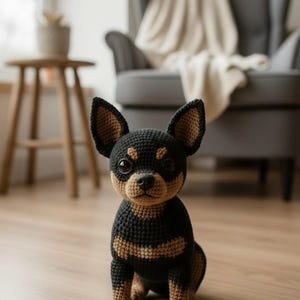 May include: A crocheted black and tan Chihuahua dog with large, dark eyes and a detailed snout. The handmade plush toy is sitting on a wooden floor. The dog's ears and paws are tan, and the body is black with tan markings.