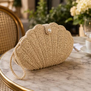 May include: A beige crochet clutch bag in the shape of a scallop shell, with a pearl button clasp. The bag has a braided handle and is on a marble table. The background features a chair and plants.