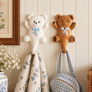 May include: Two ceramic wall hooks shaped like teddy bears. One bear is white with a light blue bow, and the other is brown with a light blue bow. Each bear has a hook at the bottom. A floral patterned towel hangs from the white bear, and a blue and white checkered bag hangs from the brown bear.