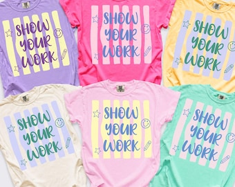 May include: Six short-sleeve crew neck t-shirts in purple, hot pink, yellow, cream, light pink, and mint green. Each shirt features a central graphic with vertical stripes, the text "SHOW YOUR WORK" in blue, and small star, smiley face, and pencil icons.