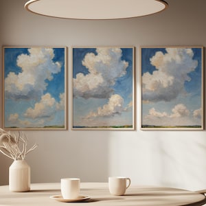 May include: Three framed cloudscape paintings with blue skies and fluffy white clouds hang above a wooden table. A vase with dried flowers and two cups and a saucer are on the table. The room has a neutral color palette.