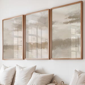 May include: Three framed abstract art prints with a neutral color palette hang above a wooden bench with white pillows. A woven basket and a striped pillow add texture to the scene. The artwork features muted tones of beige, brown, and gray.