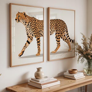 May include: Two framed art prints featuring a cheetah illustration. The cheetah is depicted in a walking pose with a tan coat and black spots. The frames are made of light-colored wood, and the background is a neutral beige.