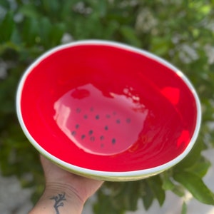 May include: A red and green ceramic bowl with a watermelon design. The bowl has a white rim and black seeds painted on the inside.