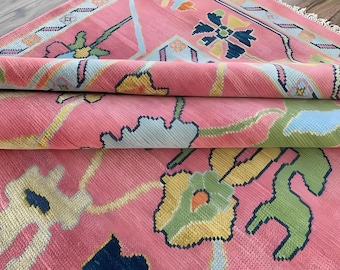 Könnte beinhalten: Eine aufgerollte rosa Teppich mit einem detaillierten Blumenmuster in Blau-, Grün-, Gelb- und Orangetönen. Der Teppich hat eine strukturierte Oberfläche und einen Fransenrand. Das Design zeigt geometrische Formen und stilisierte Blumen.
