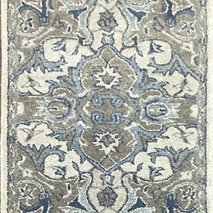 May include: A rectangular rug with an intricate, symmetrical pattern. The design features floral and geometric elements in shades of beige, brown, and blue. The rug has a cream-colored border and a detailed central motif.
