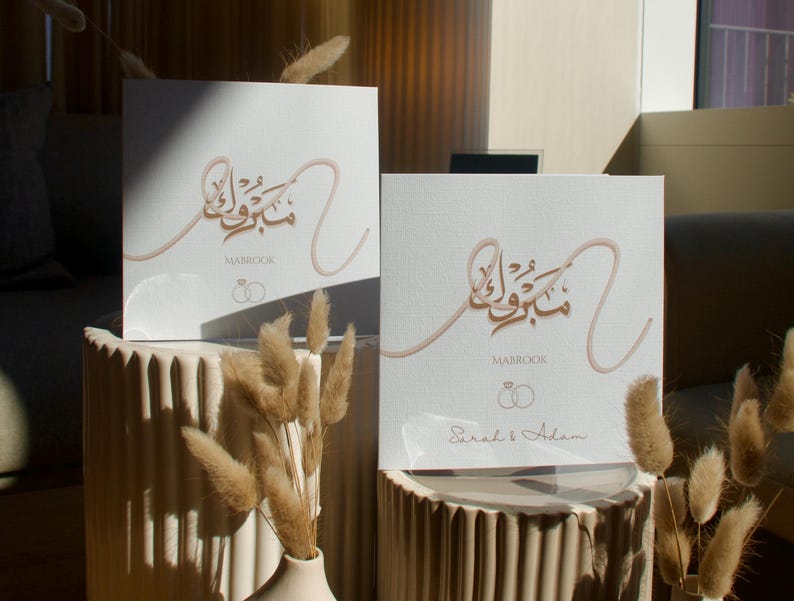 May include: Two white wedding guest books with gold calligraphy and the word "Mabrook". The books are on a beige stand with dried flowers in a vase. The names "Sarah & Adam" are printed on one book.