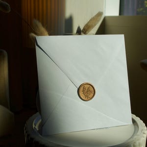 Może przedstawiać: Biała kwadratowa koperta zapieczętowana okrągłą, złotą pieczęcią woskową z motywem liścia. Koperta spoczywa na białej powierzchni, z widokiem na krzesło w tle. Scena jest oświetlona naturalnym światłem.