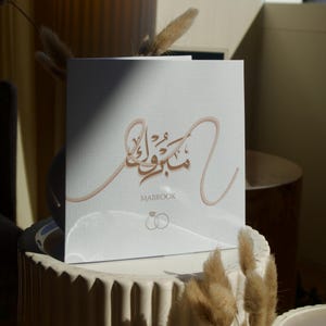 May include: A white square card with elegant calligraphy in a warm brown hue. The word "MABROOK" is printed below the calligraphy, with two intertwined rings. The card is placed on a white, textured surface, with dried plants in the foreground.