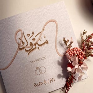 Może przedstawiać: Teksturowana, kremowa karta zaproszenia ślubnego z arabską kaligrafią i słowem "MABROOK" wydrukowanym na brązowo. Karta zawiera dwa splecione pierścienie i tekst arabski. Po prawej stronie znajduje się mała kompozycja kwiatowa z czerwonymi i r&oacute;żowymi kwiatami.