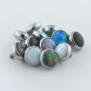 May include: A collection of body jewelry, including dermal anchors and tops. The tops feature iridescent opal-like stones in various colors, including blue, green, and purple, set in silver-colored metal. The jewelry is scattered on a white surface.