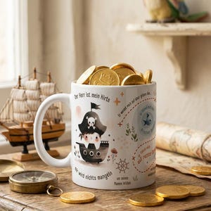 Puede incluir: Una taza de cerámica blanca llena de monedas de chocolate doradas, con una ilustración de un pirata, un barco y texto en alemán que incluye "Der Herr ist mein Hirte" y "mir wird nichts mangeln" impreso en tinta negra y marrón.