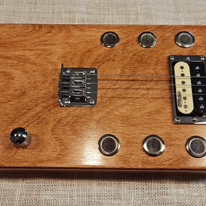 May include: A handmade wooden cigar box guitar with a natural finish. It features a fretboard, a pickup, chrome hardware, and tuning knobs. The instrument has a unique, rustic aesthetic.