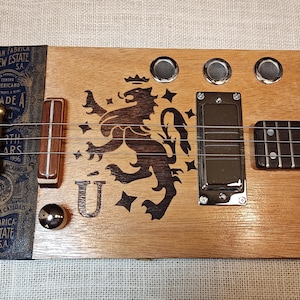 May include: A handmade cigar box guitar with a wooden body. The guitar features a black and brown lion crest, metal hardware, and strings. The headstock has the text "GRAN FABRICA DREW ESTATE S.A." and "GRADE A".