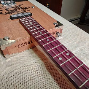 May include: A handmade cigar box guitar with a wooden body and a purple fretboard. The guitar features metal hardware, strings, and a decorative design on the body. The text "FERAL" is visible on the side of the box.