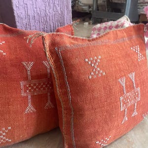May include: Two orange decorative throw pillows with white embroidered geometric patterns. The pillows have a textured appearance and are likely made of woven fabric. The design includes a central rectangular shape with vertical and horizontal lines.