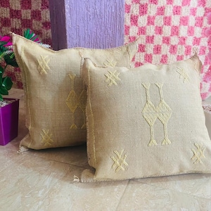 Puede incluir: Dos cojines cuadrados beige con patrones geométricos amarillos bordados. Los cojines están colocados sobre una superficie clara, con flores rosas y moradas en el fondo. Los cojines tienen un aspecto texturizado.