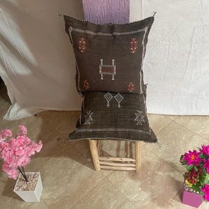 Puede incluir: Dos cojines decorativos marrones con patrones geométricos, apilados sobre un pequeño taburete de madera. Los cojines tienen detalles en blanco y rojo. El taburete es de madera clara. El fondo es una pared blanca y una pared morada texturizada.