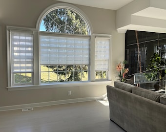 May include: A well-lit living room with large windows fitted with white, layered blinds. The windows provide a view of the outside. A grey sofa is in the foreground, and the room has light-coloured walls and flooring.