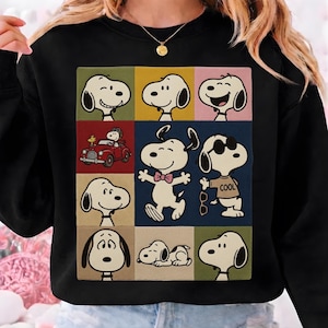 May include: Black sweatshirt featuring a colorful grid of Snoopy images. The design includes various Snoopy expressions and poses, including one driving a red car and another wearing sunglasses. The word "COOL" is printed on one of the Snoopy's shirts.