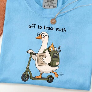 May include: A light blue t-shirt featuring a graphic of a white goose wearing glasses, riding a green scooter, and carrying a green backpack filled with math supplies. The goose holds a book labeled MATH with a triangle and the text y=mx+b. The text off to teach math is printed above the goose.