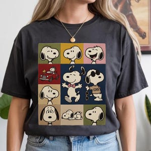 May include: Dark gray t-shirt featuring a colorful grid of Snoopy images. The grid includes Snoopy in various poses, including driving a red car, wearing sunglasses, and with a bow tie. A gold necklace is visible.