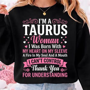 May include: Black sweatshirt with the text "I'M A TAURUS Woman" in white and pink. The design includes hearts and floral accents, with the phrases "I Was Born With My Heart On My Sleeve" and "Thank You For Understanding".