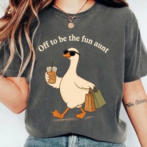 May include: A charcoal grey t-shirt featuring a graphic of a white goose wearing black sunglasses, holding a brown iced coffee cup in one wing and carrying two shopping bags in the other, with the text "Off to be the fun aunt" printed above.