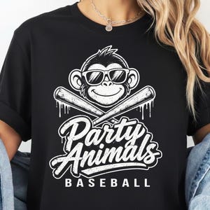 May include: A black t-shirt featuring a white graphic of a monkey wearing sunglasses above two crossed baseball bats. Below the bats, the text reads Party Animals in a stylized script font, with the word BASEBALL printed in capital letters underneath.