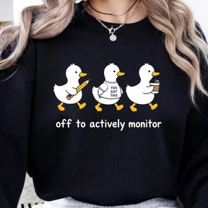 May include: Black sweatshirt featuring three cartoon ducks. The first duck holds a pencil, the second wears a shirt that says "You Got This," and the third carries a coffee cup. The text "off to actively monitor" is printed below the ducks.