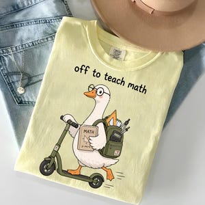 May include: A pale yellow t-shirt featuring a graphic of a white goose wearing glasses, riding a green scooter, and carrying a green backpack filled with school supplies. The text "off to teach math" is printed above the goose in black.