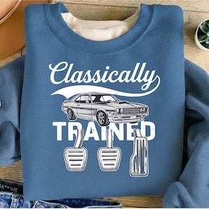 Może przedstawiać: Niebieska bluza z białym napisem "Classically Trained", ilustracją klasycznego samochodu i trzema pedałami. Bluza ma okrągły dekolt i miękki, wygodny wygląd.