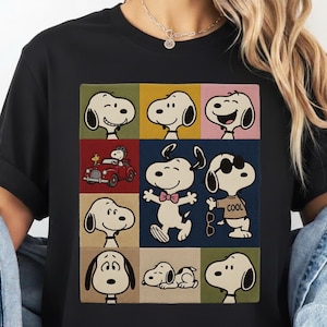 May include: Black t-shirt featuring a colorful grid of Snoopy images in various poses. The design includes Snoopy in a red car, wearing sunglasses, and with a bow tie. The shirt is made of a soft material.
