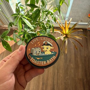 May include: A round patch with the words "LIVE, LAUGH" at the top and "TOASTER BATH" at the bottom. The patch features a cartoon toaster and a goat in a bathtub. The patch is brown, black, and tan.