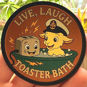 May include: A round, brown and green patch with the words "LIVE, LAUGH, TOASTER BATH". The patch features a cartoon toaster and a goat in a bathtub, both with smiling faces. The goat wears a navy hat with a gold emblem.
