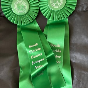 May include: Two green rosettes with matching ribbons. Each rosette has a circular emblem with text and a horse and rider silhouette. The ribbons read "South Florida Hunter Jumper Assn."