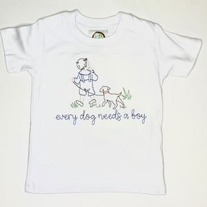 Op de afbeelding: Wit T-shirt met een geborduurd ontwerp van een kind dat een hond aan de lijn uitlaat. De tekst "every dog needs a boy" is eronder in blauwe letters geborduurd. Het shirt heeft korte mouwen en een ronde hals.