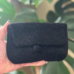 May include: A small, rectangular black beaded clutch with a flap closure and a round button clasp. The clutch is held in a hand, with a blurred green leafy background.